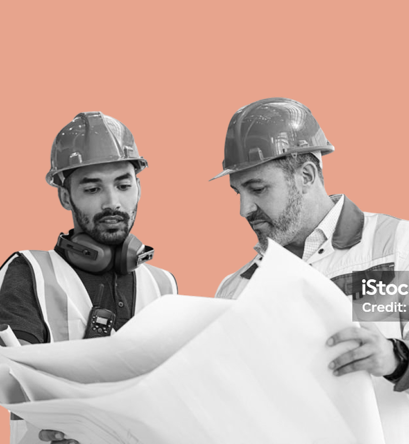Two construction workers wearing hard hats and safety vests, closely examining large blueprints on a solid peach background.