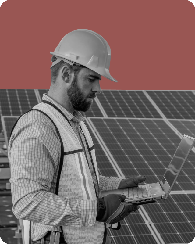 A male engineer wearing a hard hat, safety vest, and gloves, working on a laptop in front of large solar panels.