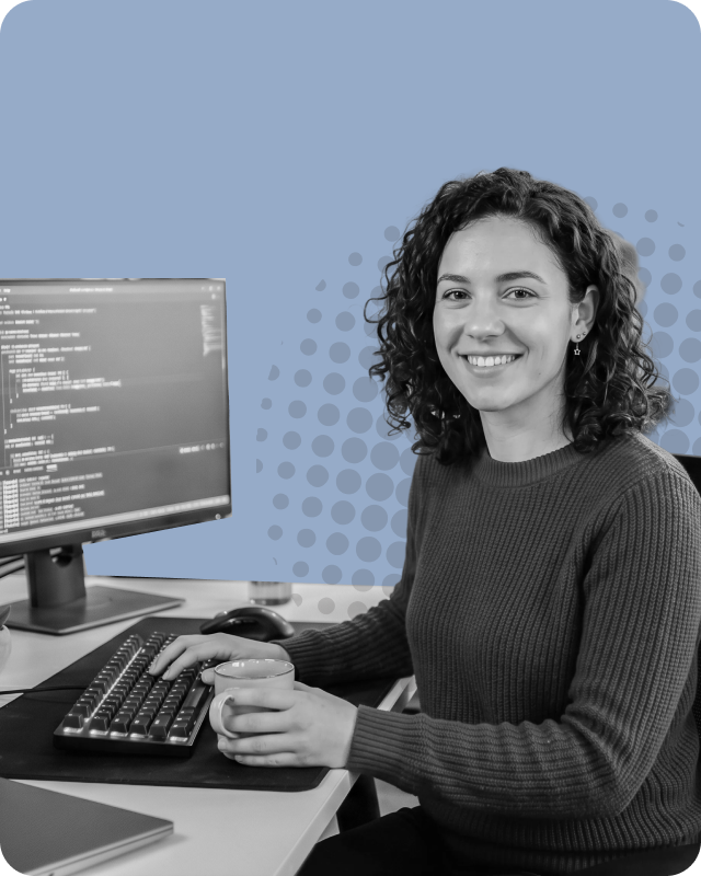 Smiling woman with curly hair sitting at a desk working on a computer with code displayed on the monitor, holding a coffee cup.