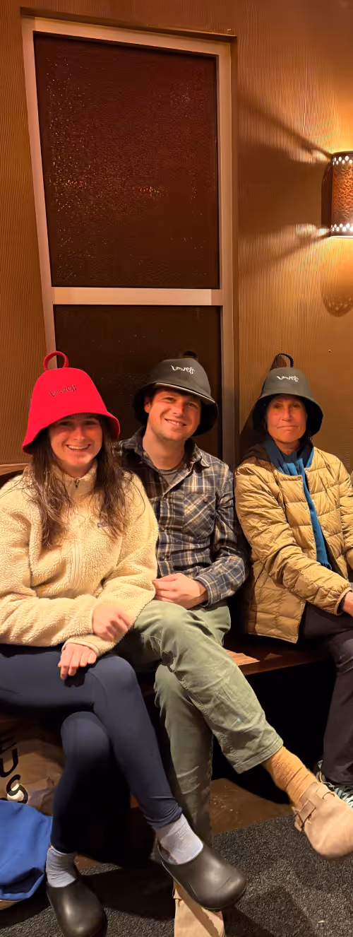 Three people sitting on a bench indoors, wearing bucket hats; one in red and two in black, smiling at the camera.