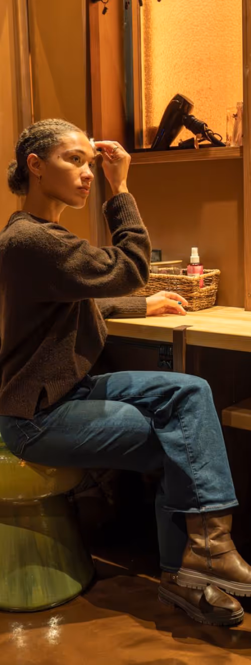 Person with curly hair sitting on a green stool applying makeup in front of a wooden vanity with a basket and hairdryer.