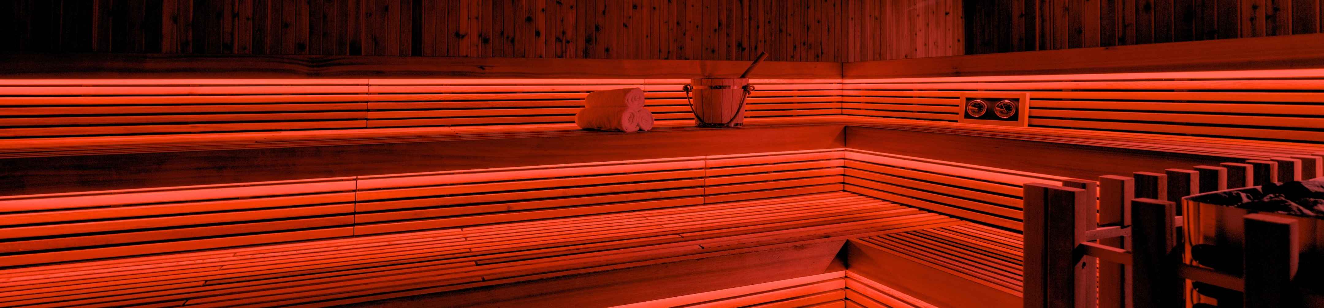 Interior of a wooden sauna with orange ambient lighting, towels, and a water bucket on the bench.