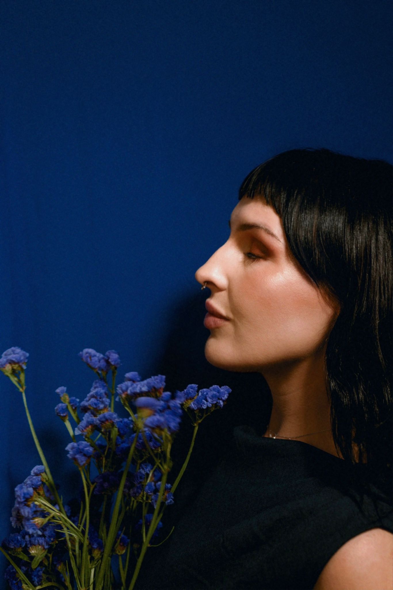 A woman with long dark hair and a septum piercing stands in profile against a deep blue background, holding a bouquet of small purple flowers. Her eyes are closed and she has a calm, serene expression