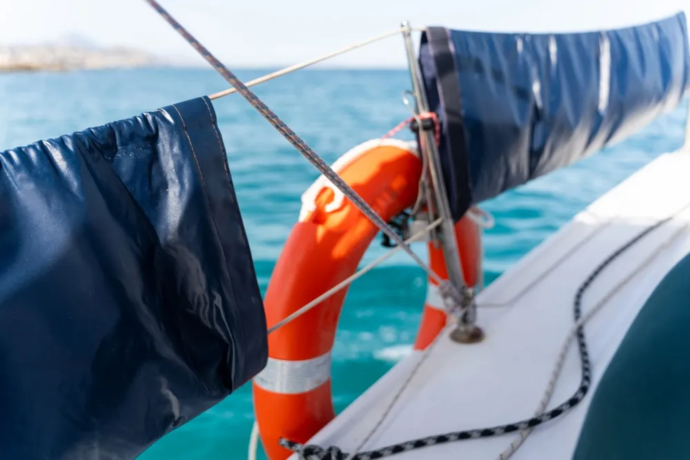 Close-up of a sailboat deck featuring a bright orange lifebuoy tied with ropes. Blue sea and distant shorelines are visible in the blurred background, conveying a serene nautical scene.