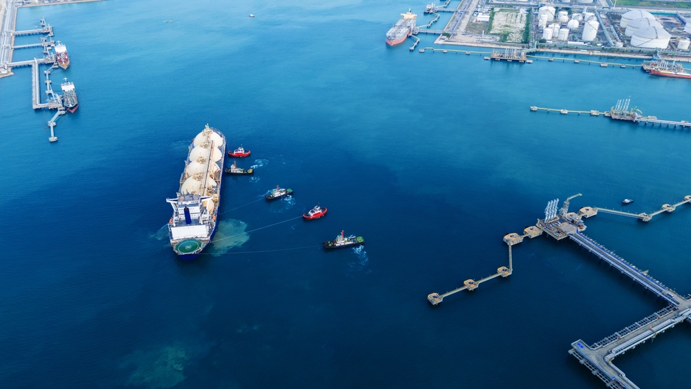 Aerial view of a busy harbor with a large cargo ship flanked by tugboats in deep blue water. Nearby docks and storage tanks indicate a bustling port.