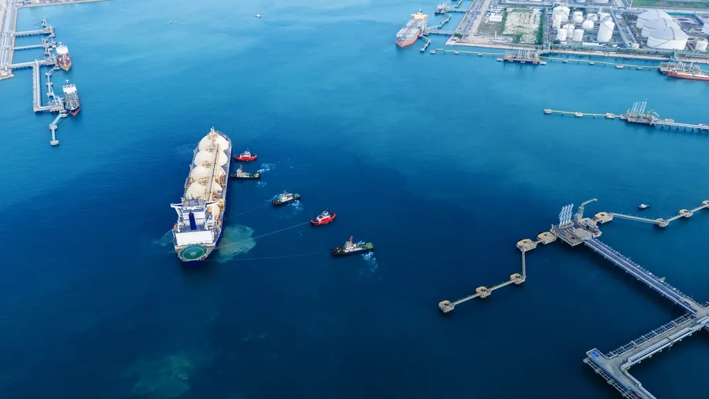 Aerial view of a busy harbor with a large cargo ship flanked by tugboats in deep blue water. Nearby docks and storage tanks indicate a bustling port.