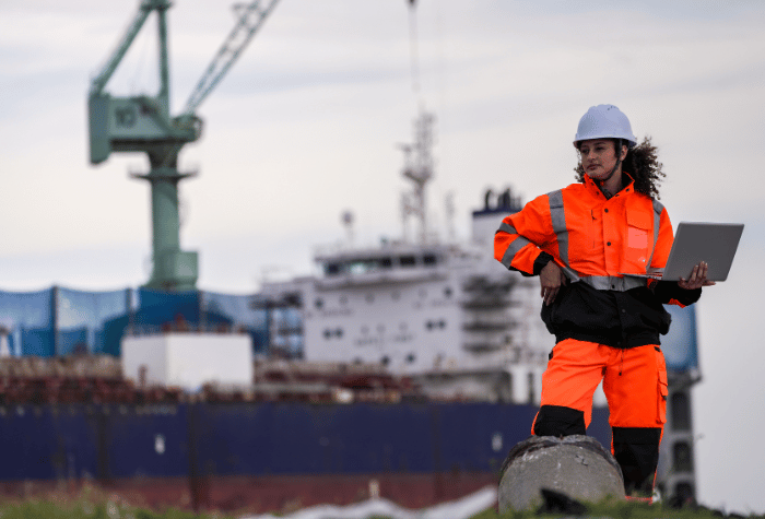 Maritime worker in orange jumpsuit holds laptop in front of container ship