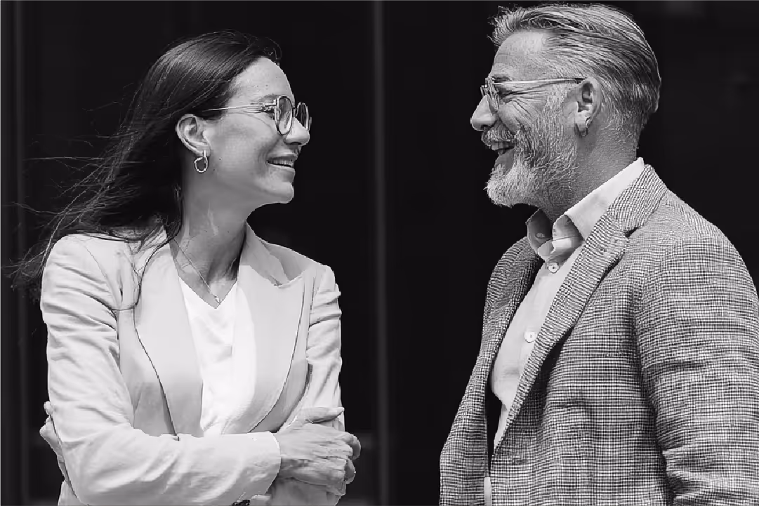 Black and white photo of a smiling middle-aged man with glasses and beard talking to a smiling woman with glasses and long hair.