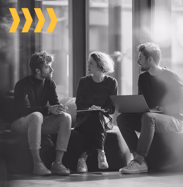 Three young professionals seated indoors discussing, one with a laptop, with four yellow arrow icons above them.
