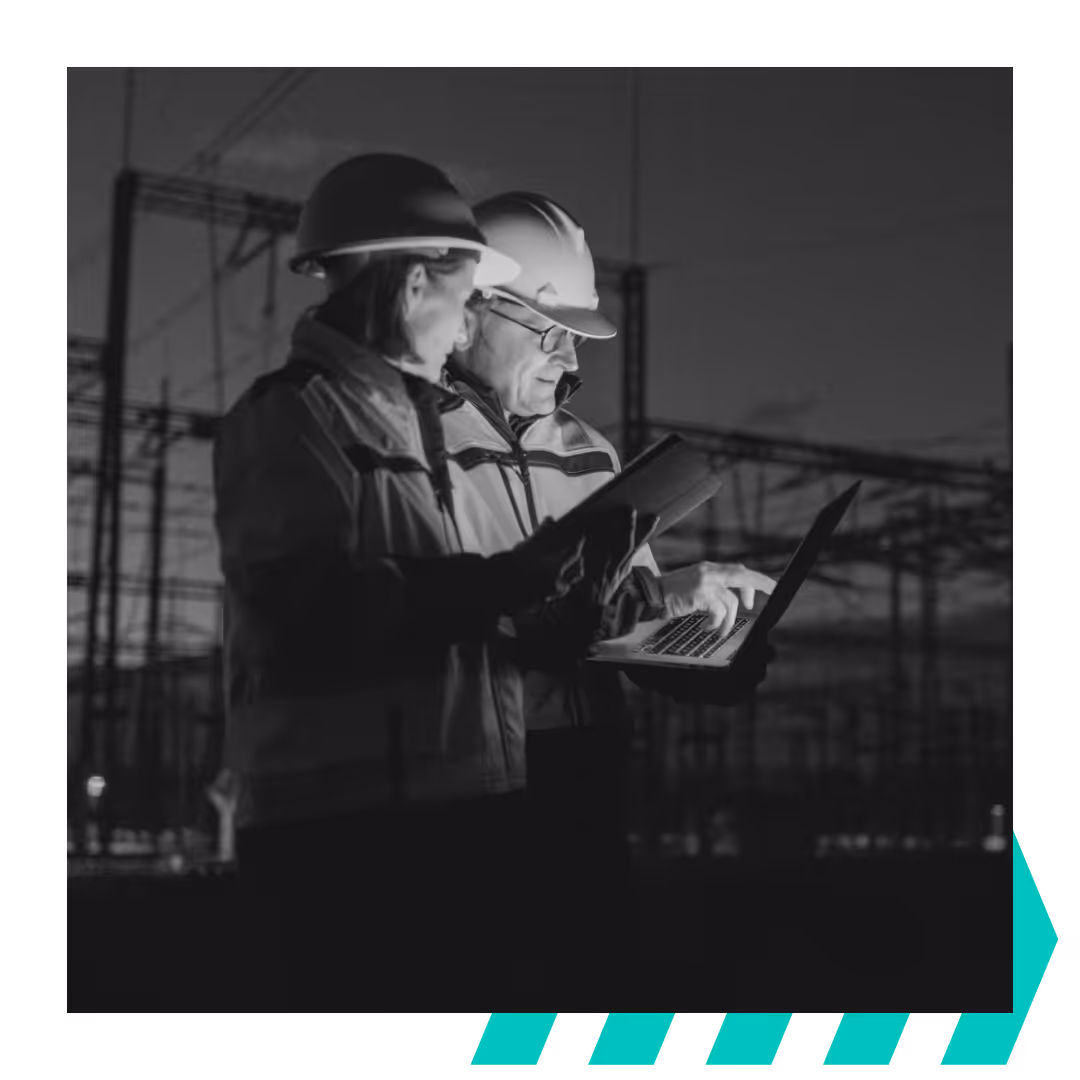 Two construction workers wearing helmets reviewing information on a tablet and laptop at an electrical substation during dusk.