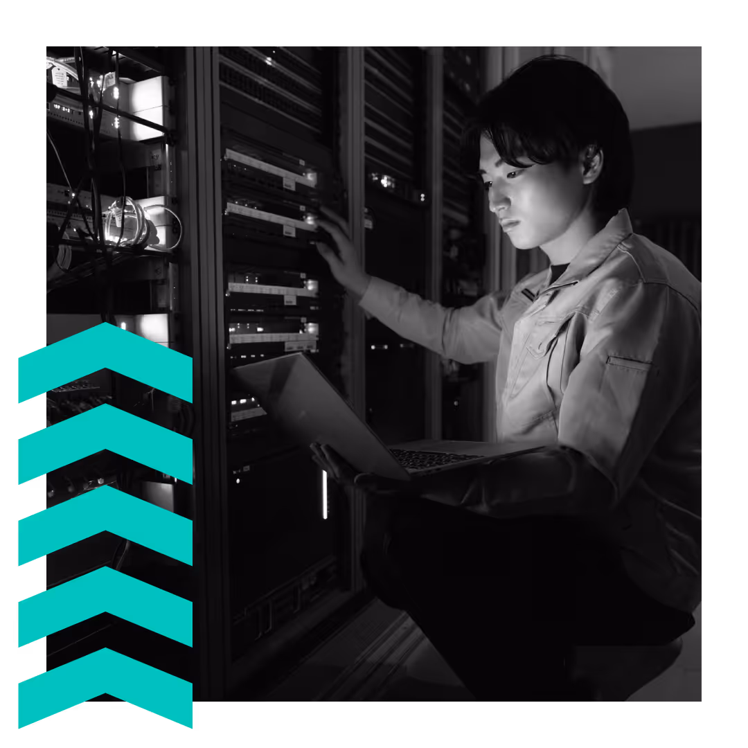 Technician working on server racks while using a laptop in a dimly lit data center.