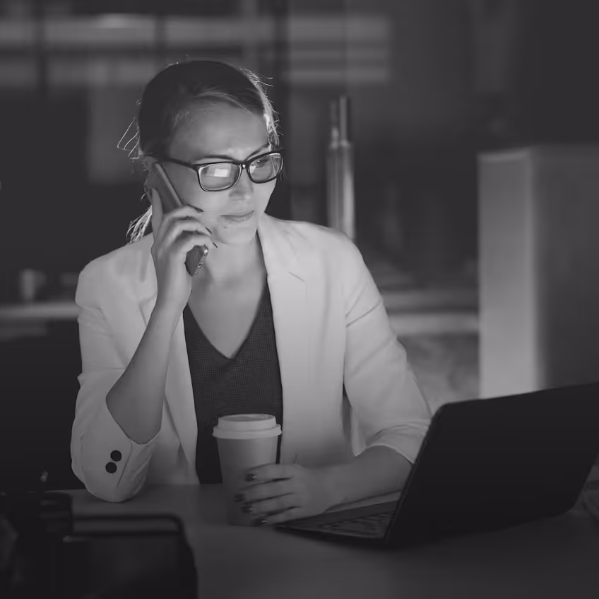 Woman wearing glasses and white blazer talking on a smartphone while sitting at a table with a laptop and holding a coffee cup.