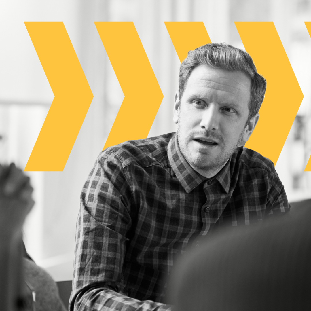 Man in a plaid shirt listening attentively in a meeting with yellow arrows behind him.