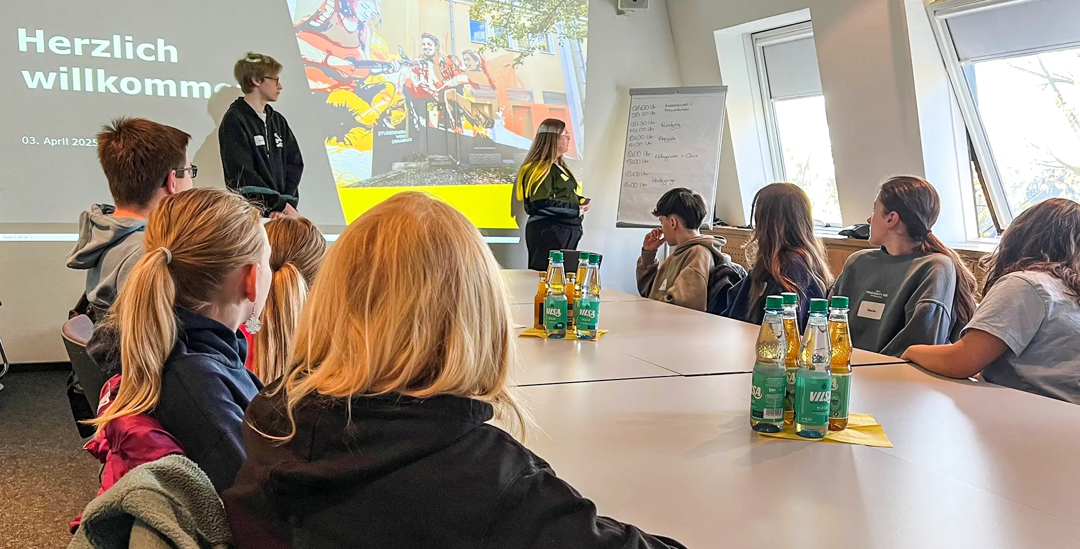 Jugendliche sitzen an einem Besprechungsraum und hören zwei Vortragenden zu, die vor einer Leinwand mit der Projektion 'Herzlich willkommen' stehen.