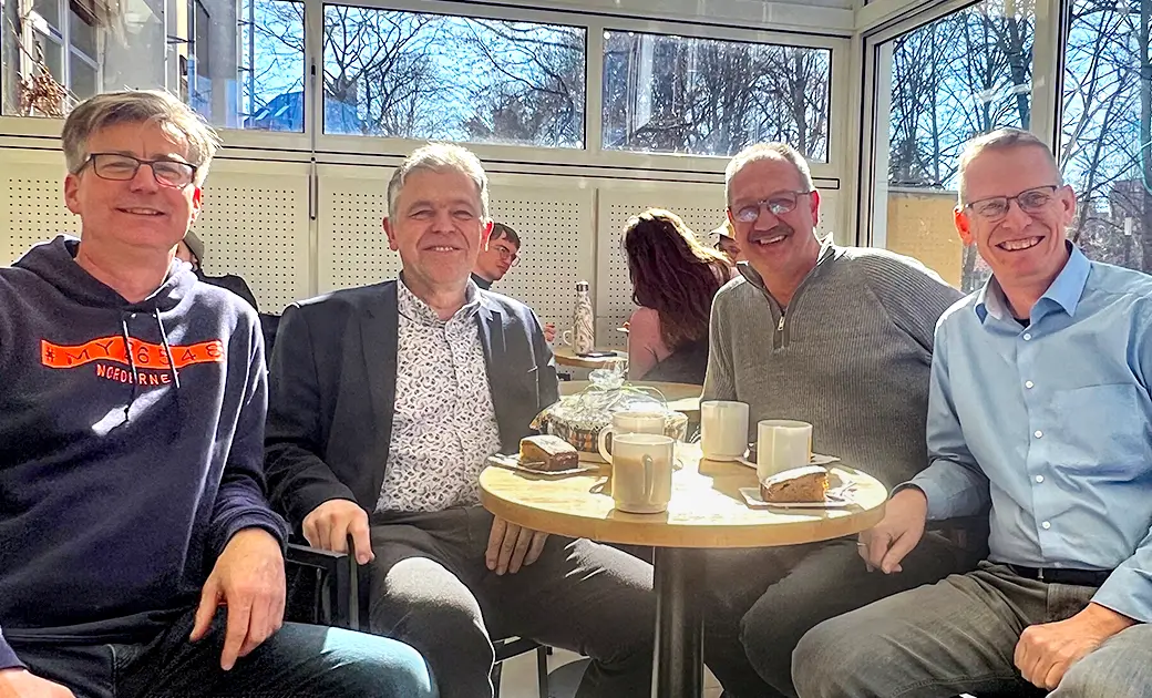 Vier Männer sitzen an einem runden Tisch mit Tassen und Kuchenstücken in einem hellen Raum eines Cafes mit großen Fenstern.