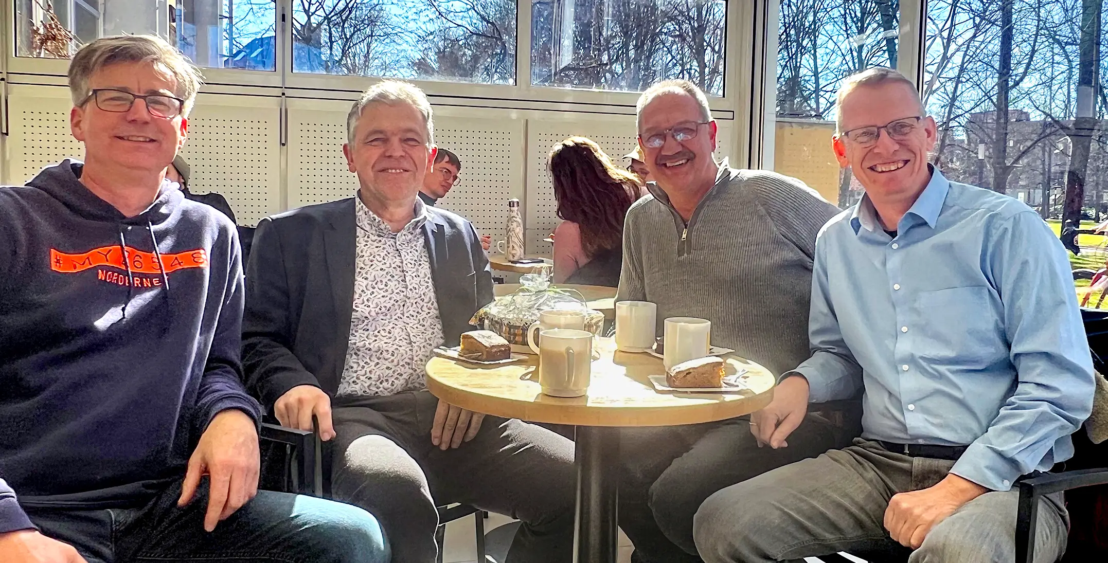 Vier Männer sitzen an einem runden Tisch mit Tassen und Kuchenstücken in einem hellen Raum eines Cafes mit großen Fenstern.