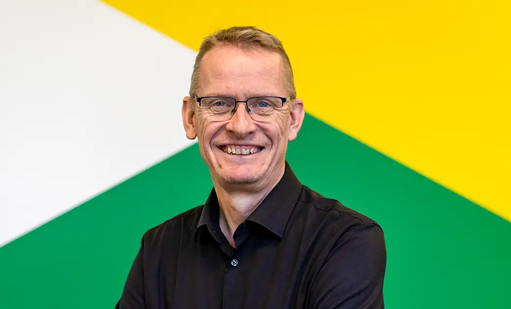 Geschäftsführer Stefan Kobilke steht vor einer Wand mit gelb-weiß-grünem geometrischem Muster und lacht freundlich in die Kamera.