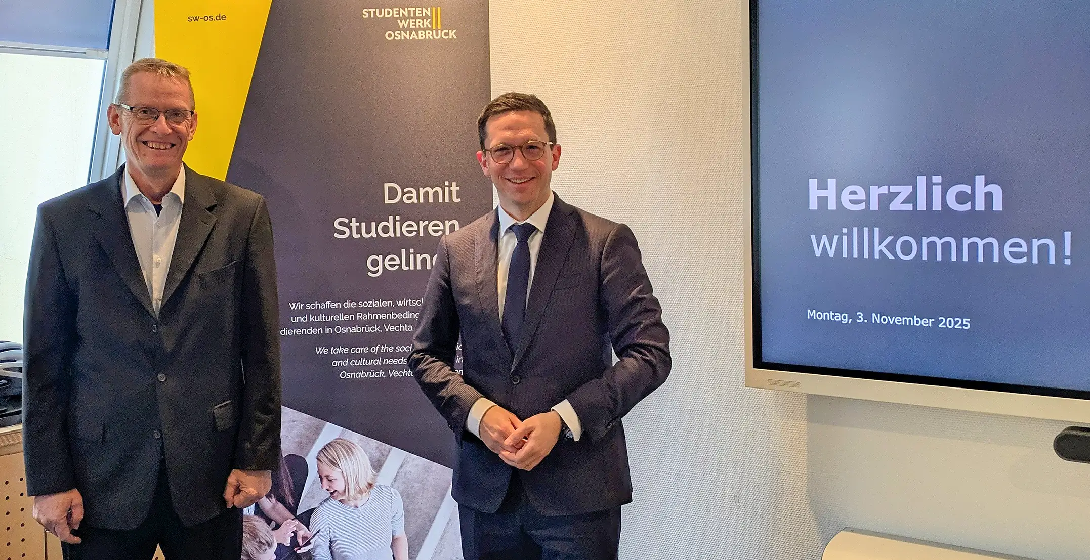 Stefan Kobilke und Falko Mohr stehen in Anzügen vor einem Banner des Studierendenwerks Osnabrück und einem Bildschirm mit der Begrüßungsnachricht 'Herzlich willkommen'.