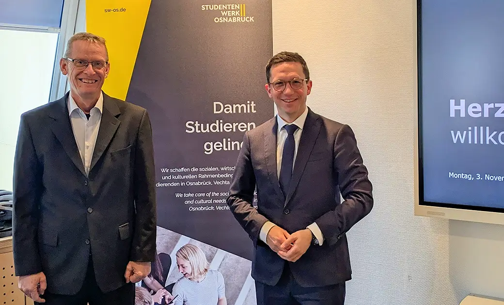 Stefan Kobilke und Falko Mohr stehen in Anzügen vor einem Banner des Studierendenwerks Osnabrück und einem Bildschirm mit der Begrüßungsnachricht 'Herzlich willkommen'.