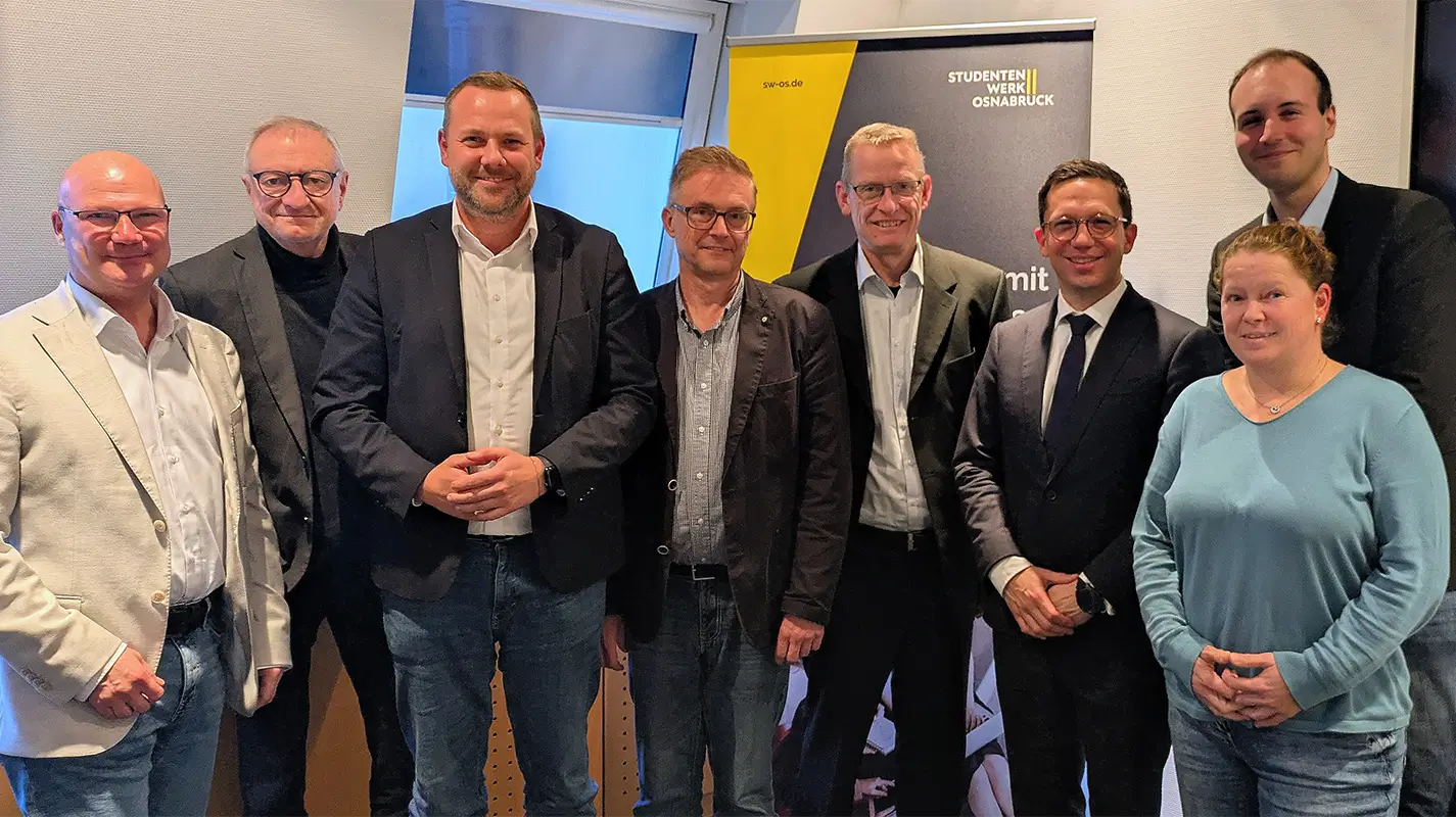 Gruppe von acht Personen steht in einem Besprechungsraum vor einem Banner mit Texten 'sw-os.de' und 'STUDIERENDENWERK OSNABRÜCK'.