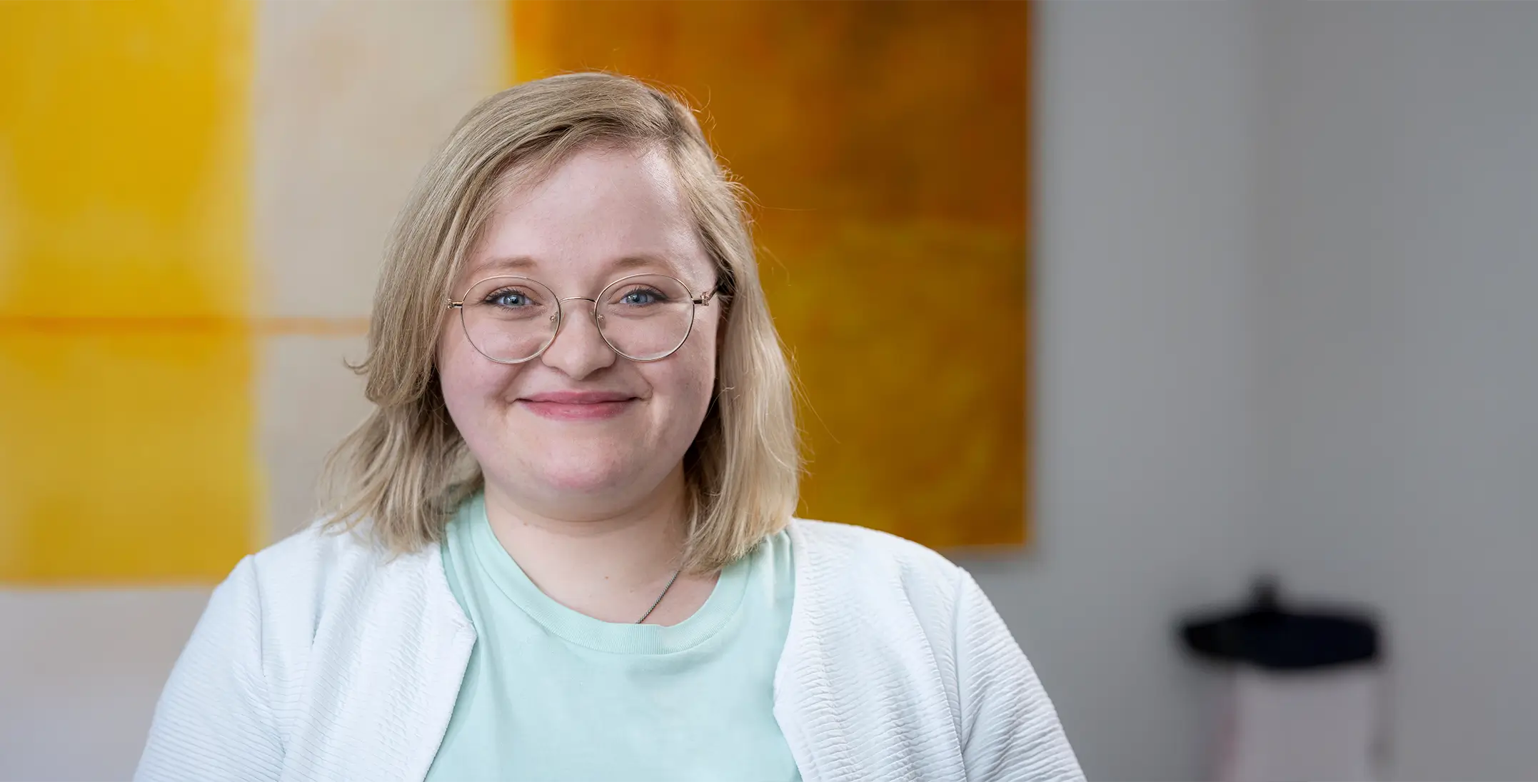 Rebekka Kalka trägt ein hellblaues T-Shirt und eine weiße Strickjacke vor einem unscharfen Hintergrund mit gelb-orangefarbenem Bild.