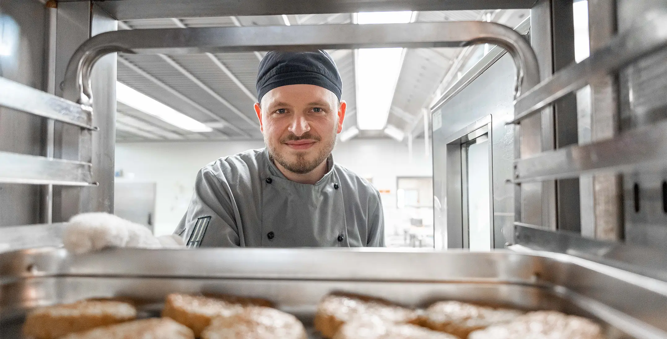 Koch in Kochjacke und Mütze blickt in einen Backofen mit mehreren Bratlingen in einer Hochschulküche.