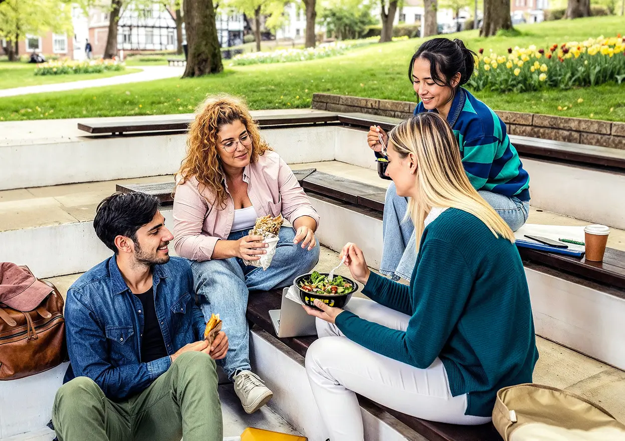 Vier Studierende sitzen im Freien in einem Park auf Stufen und essen aus Mehrwegschalen.
