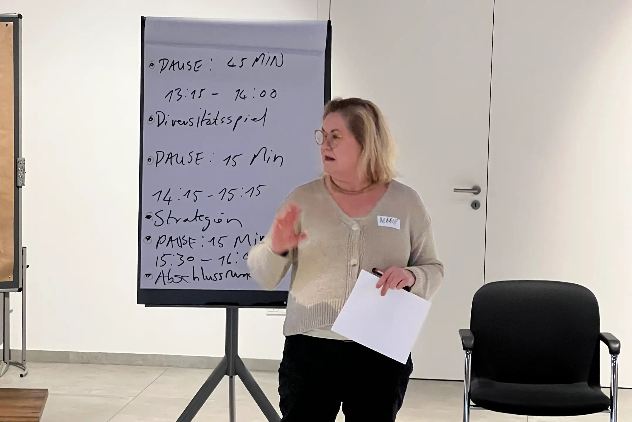 Frau steht vor Flipchart mit Zeitplan für Workshop zum Thema Diversität, hält Papier und gestikuliert.