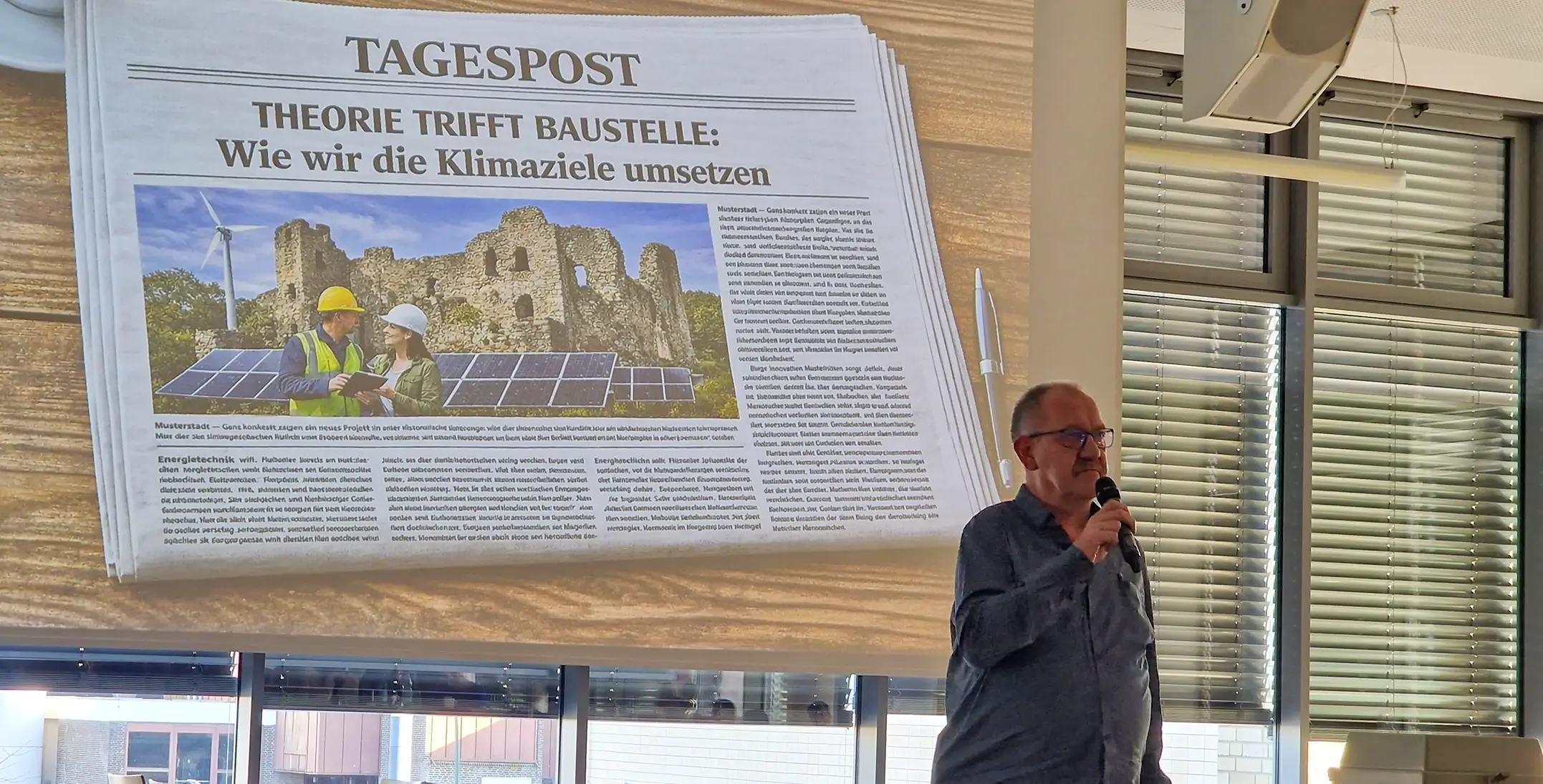 Mann mit Mikrofon vor Fenster mit Jalousien stehend neben projizierter Zeitungsseite mit Überschrift 'THEORIE TRIFFT BAUSTELLE: Wie wir die Klimaziele umsetzen' und Bild von Solarpanels vor Ruine.