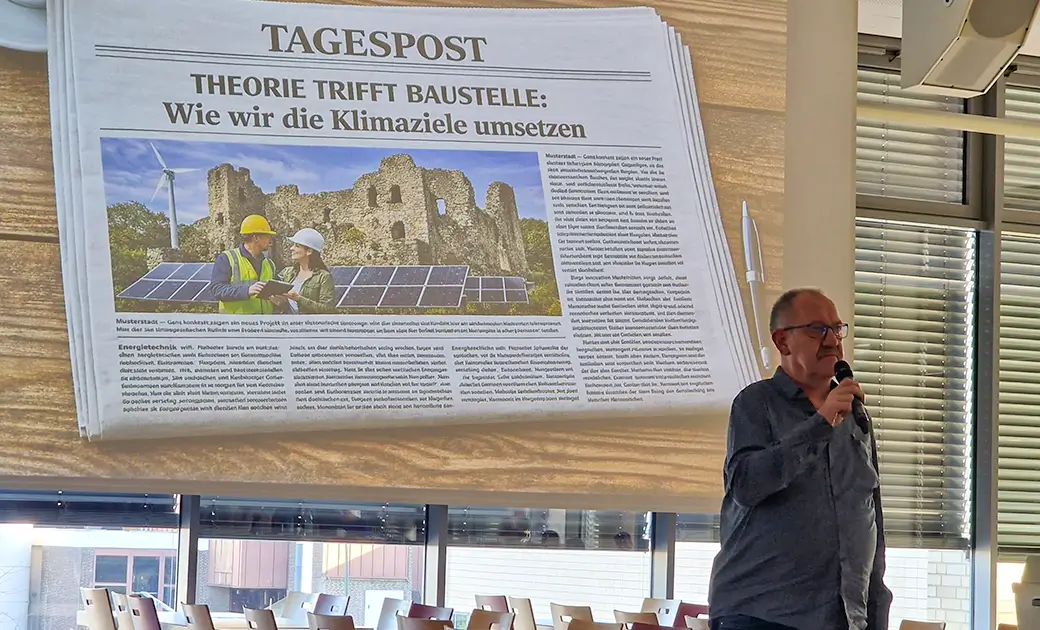 Mann mit Mikrofon vor Fenster mit Jalousien stehend neben projizierter Zeitungsseite mit Überschrift 'THEORIE TRIFFT BAUSTELLE: Wie wir die Klimaziele umsetzen' und Bild von Solarpanels vor Ruine.