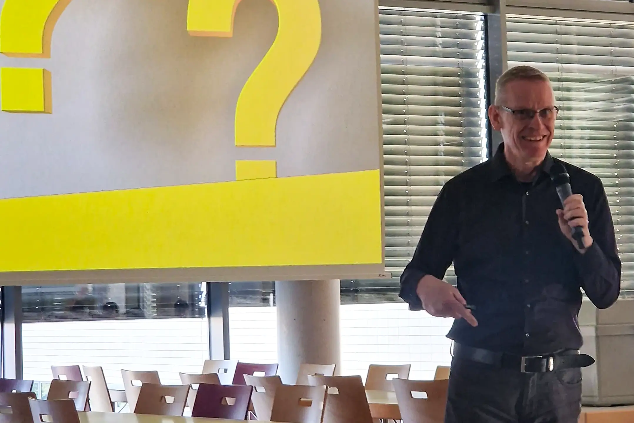 Stefan Kobilke steht mit einem Mikrofon vor einem großen gelben Fragezeichen auf einer Leinwand in einem Raum mit Stuhlreihen und Fensterjalousien.