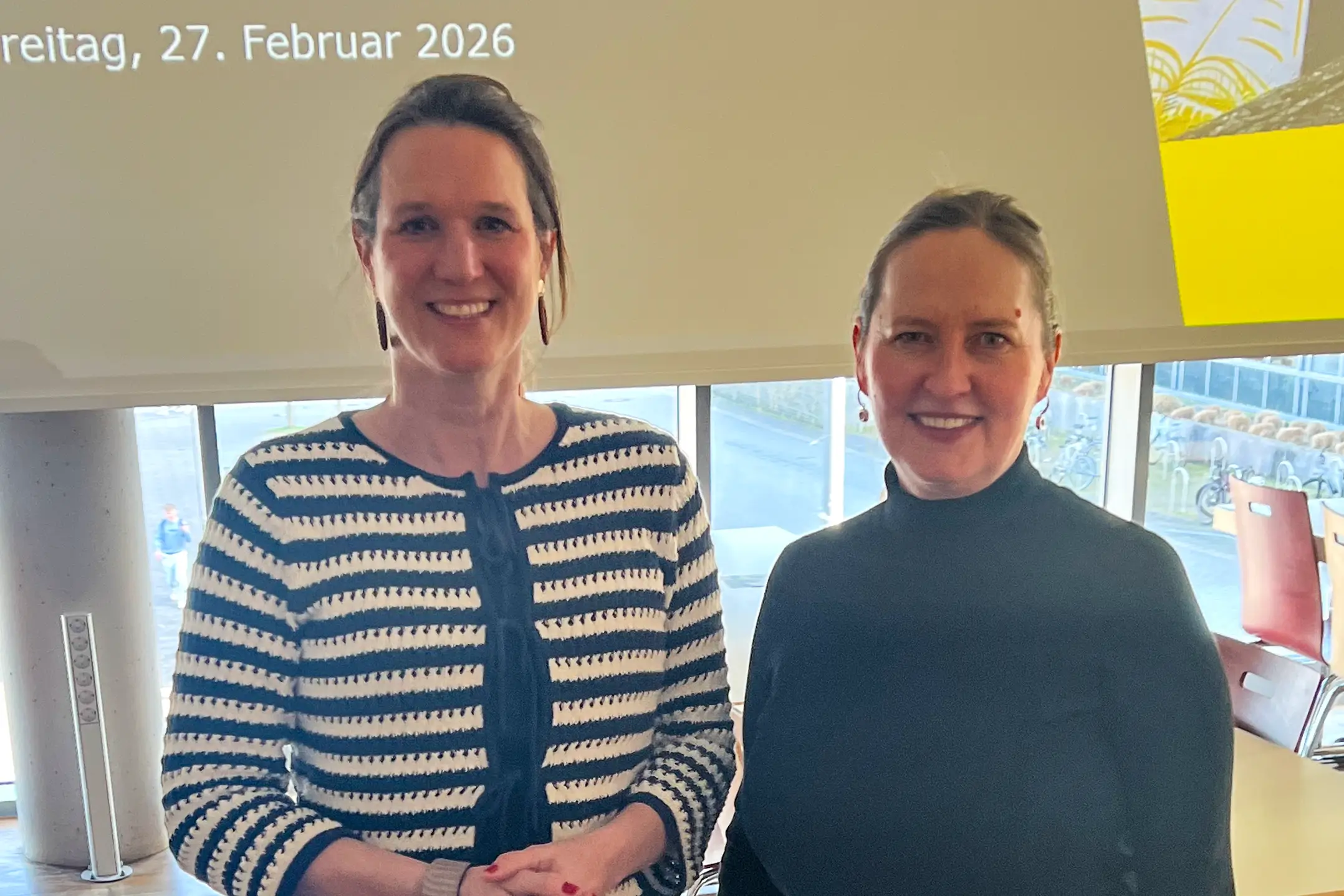 Zwei stehende Personen vor einem Projektionsbildschirm mit dem Text 'Mitarbeitendenversammlung Freitag, 27. Februar 2026' in einem hellen, modernen Raum mit Tischen und Stühlen.
