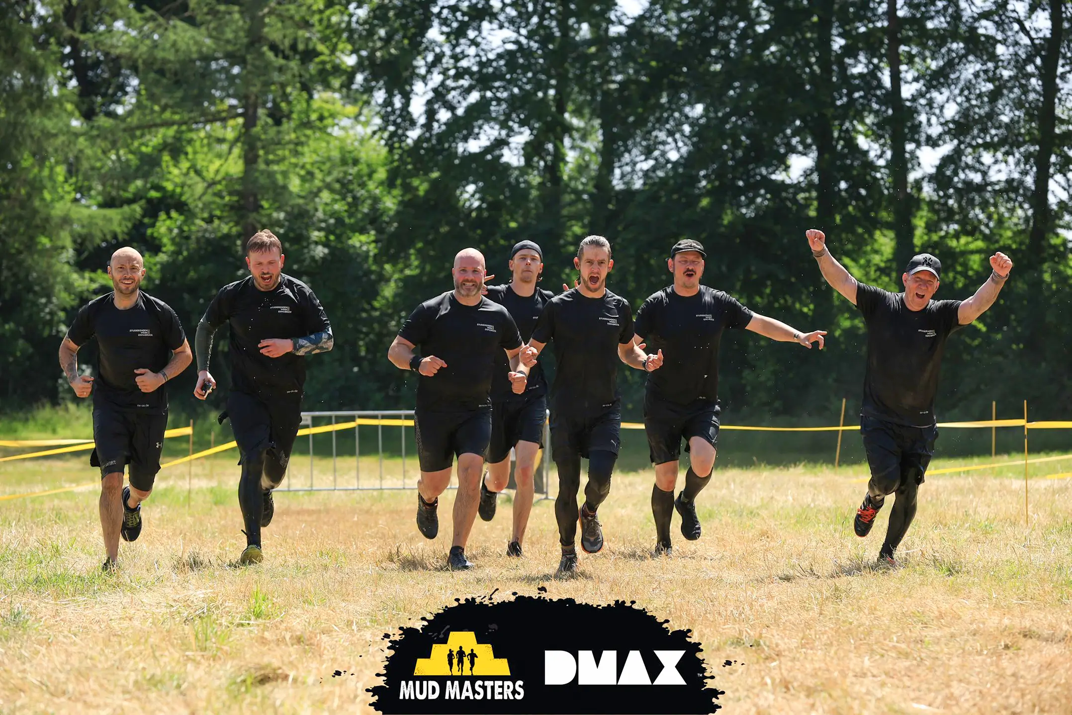Sieben Männer in schwarzer Sportkleidung laufen auf einer Wiese mit Bäumen im Hintergrund, einige mit erhobenen Armen, im Rahmen eines Mud-Muster-Teambuilding-Events.
