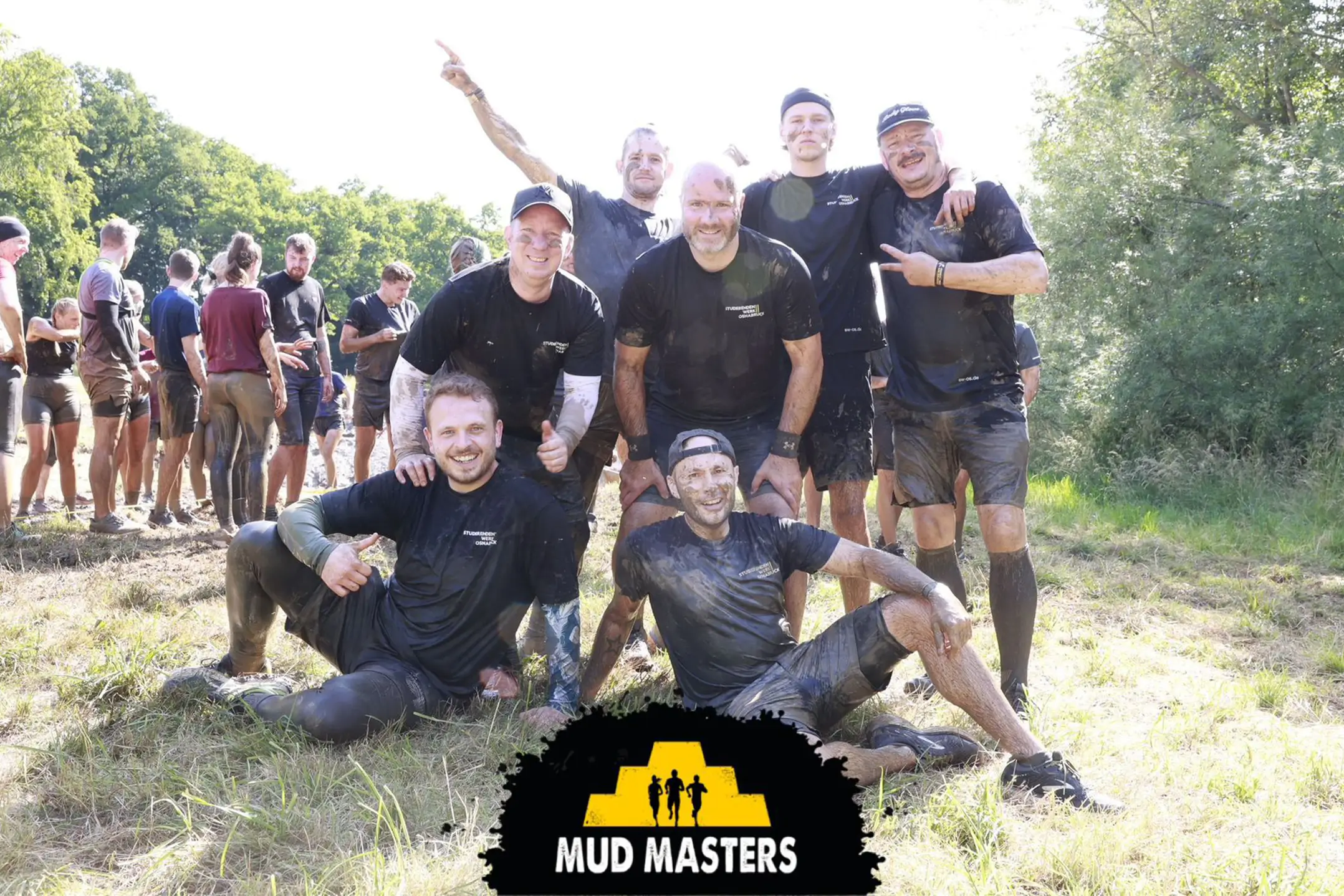 Gruppe von sieben schlammverschmierten Personen in schwarzer Sportkleidung posiert auf einer Wiese bei einem Mud-Muster-Teambuilding-Event.