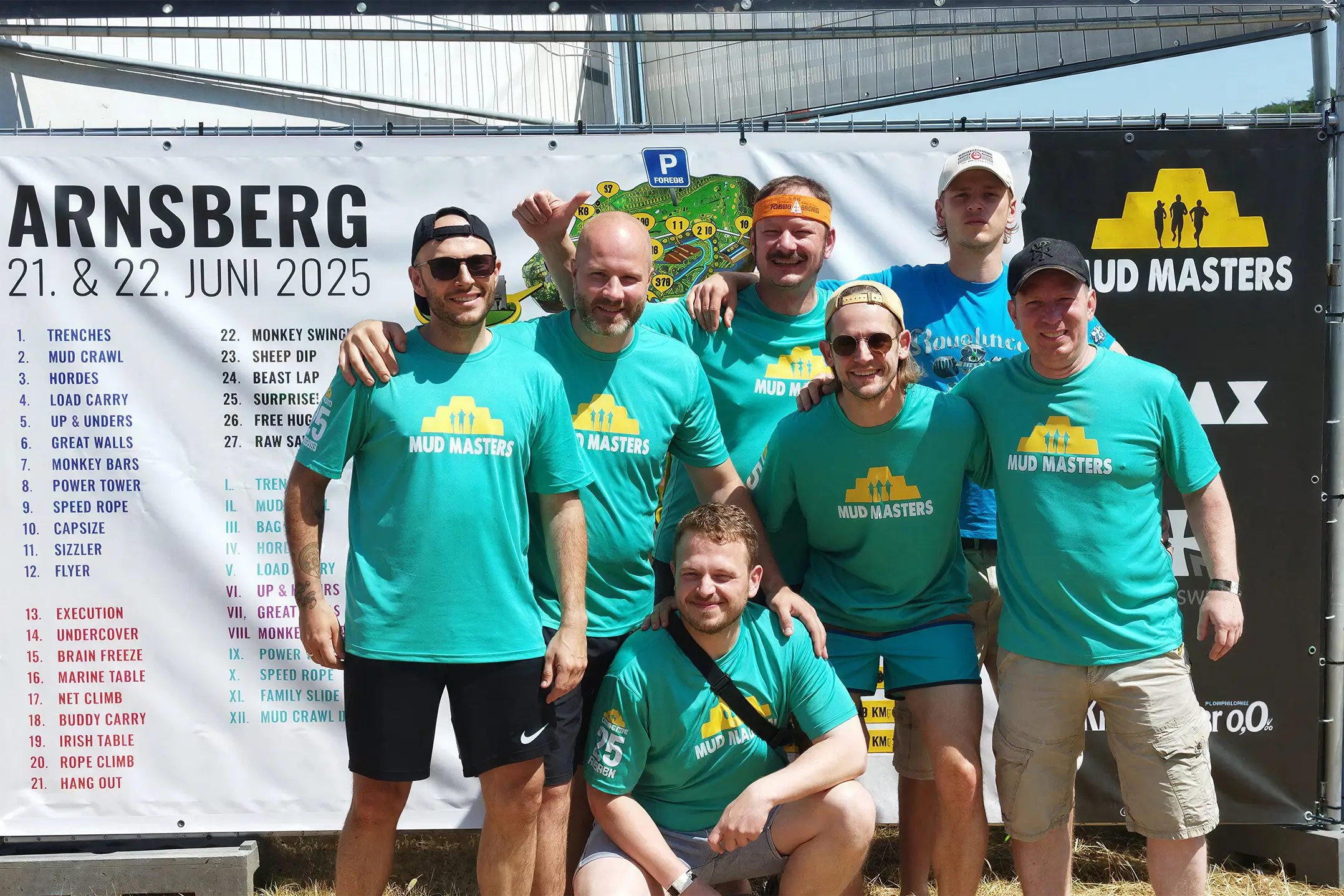 Gruppe von sieben Personen in türkisfarbenen Mud Masters T-Shirts posiert vor einem Veranstaltungsplakat für das Mud Masters Event in Arnsberg am 21. und 22. Juni 2025.