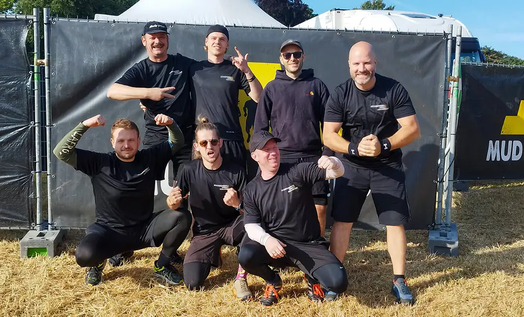 Gruppe von sieben Personen in schwarzer Sportkleidung posiert vor einem Zaun mit einem Banner, auf dem teilweise das Wort 'MUD' zu sehen ist.