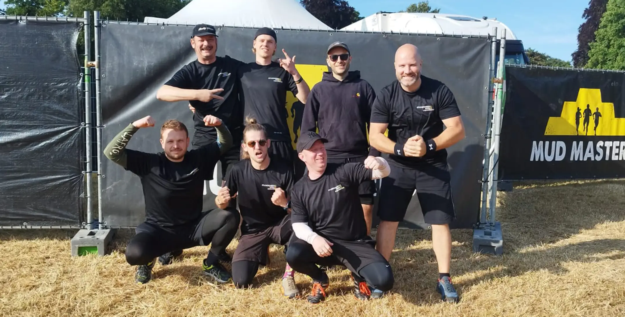 Gruppe von sieben Personen in schwarzer Sportkleidung posiert vor einem Zaun mit einem Banner, auf dem teilweise das Wort 'MUD' zu sehen ist.