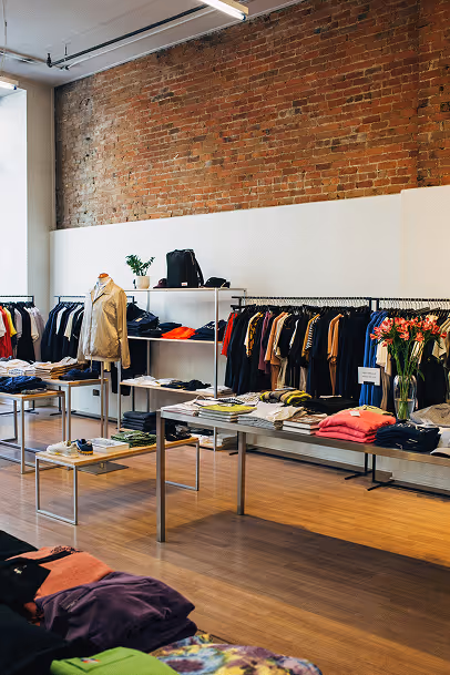 Intérieur d’une boutique de vêtements moderne avec murs en briques apparentes, portant des étagères et portants remplis de vêtements colorés, ainsi que des tables présentant des piles de vêtements soigneusement pliés. Un mannequin et un vase de fleurs apportent une touche décorative.