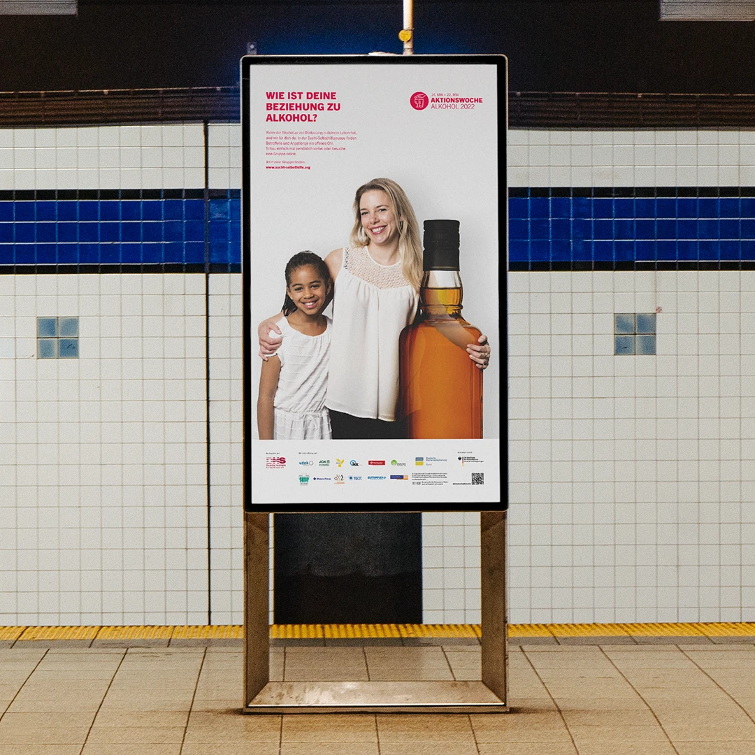 Außenplakat der Kampagne „Aktionswoche Alkohol 2024“, auf dem eine lächelnde Frau und ein Kind neben einer übergroßen Schnapsflasche stehen, mit der Überschrift „Wie ist deine Beziehung zu Alkohol?“