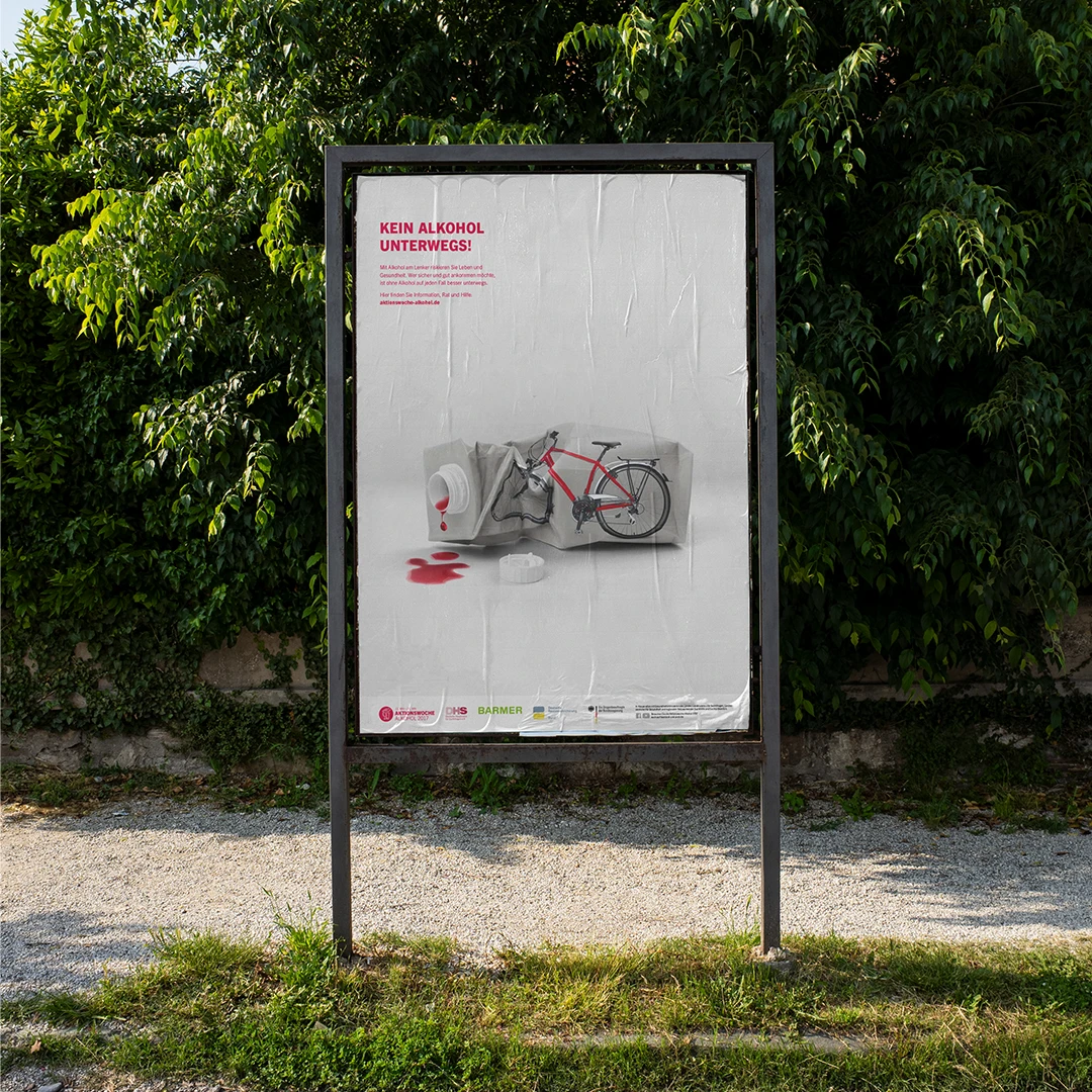 Das Bild zeigt ein Plakat an einem Straßenlaternenpfosten.  Auf dem Plakat ist eine zerstörte Gepäck abgebildet, aus der rote Flüssigkeit (vermutlich Blut) ausläuft. Ein Fahrrad ist auf der Flasche abgebildet. Der Slogan lautet „KEIN ALKOHOL UNTERWEGS!“. Das Plakat ist eine Kampagne gegen Alkohol am Steuer beziehungsweise beim Fahrradfahren.
