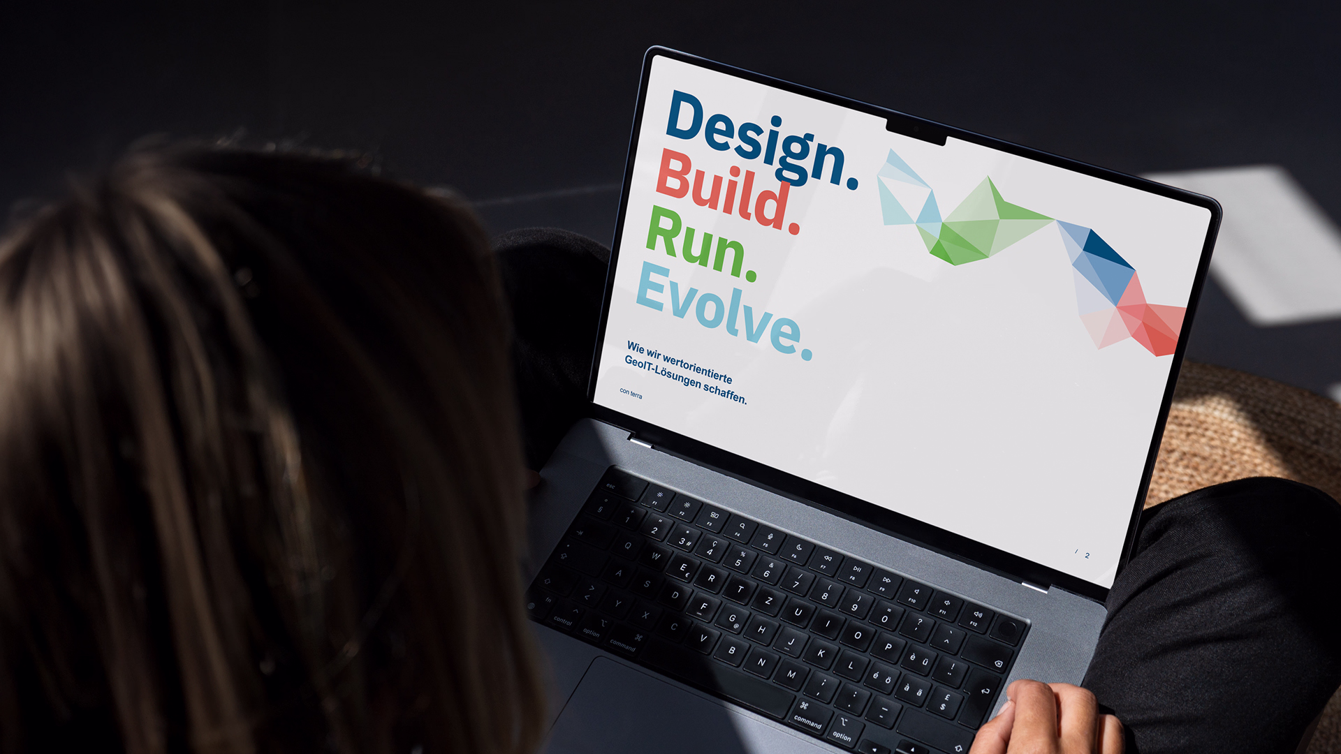 Person von hinten betrachtet, die Laptop mit Bildschirm zeigt, auf dem Text 'Design. Build. Run. Evolve.' und farbige geometrische Formen zu sehen sind.