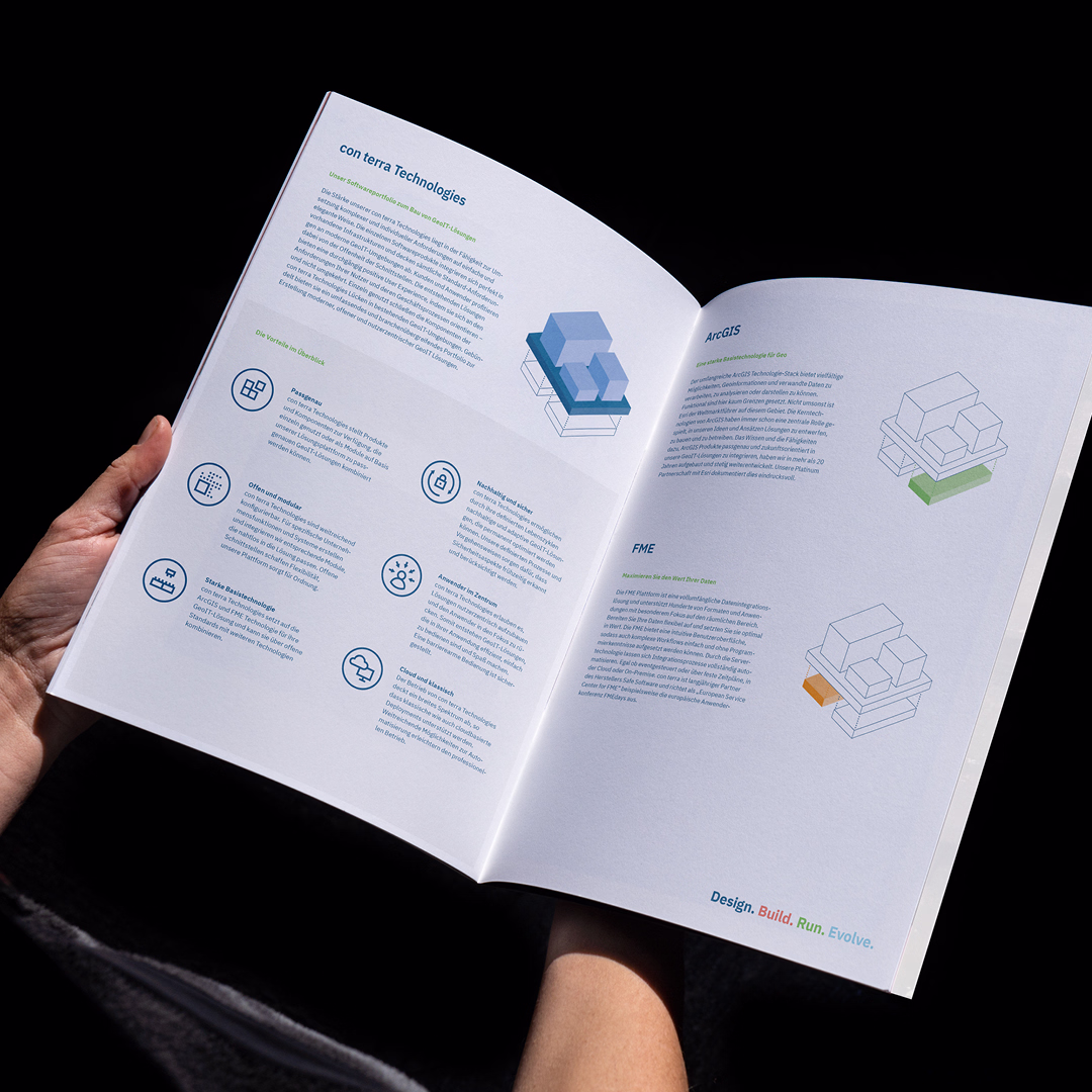 Aufgeschlagene Broschüre mit Text und schematischen 3D-Grafiken von Gebäuden, gehalten von zwei Händen vor schwarzem Hintergrund.