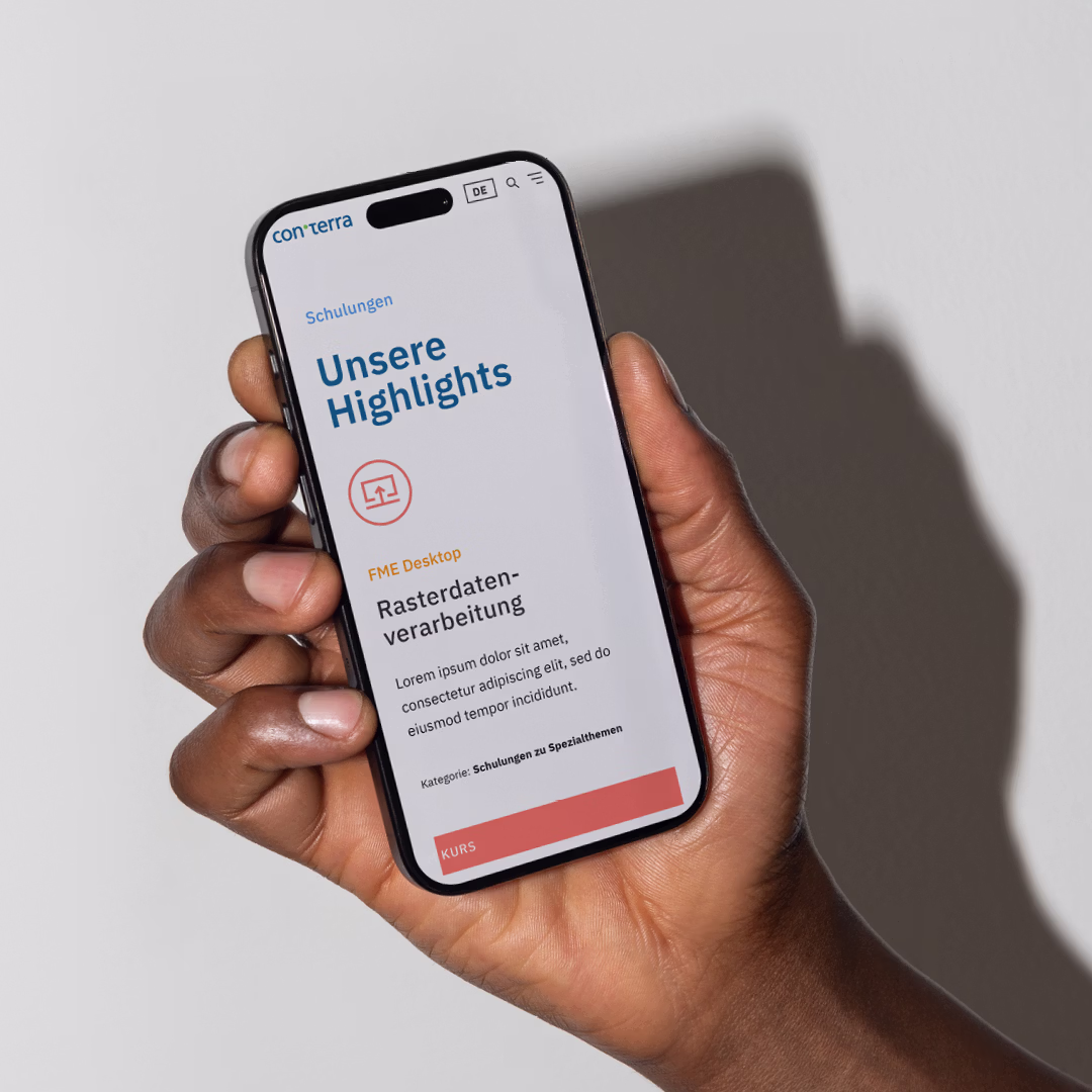 Hand hält Smartphone mit Webseite von con terra, Überschrift 'Unsere Highlights', Abschnitt 'FME Desktop Rasterdatenverarbeitung' und rotem Button 'KURS'
