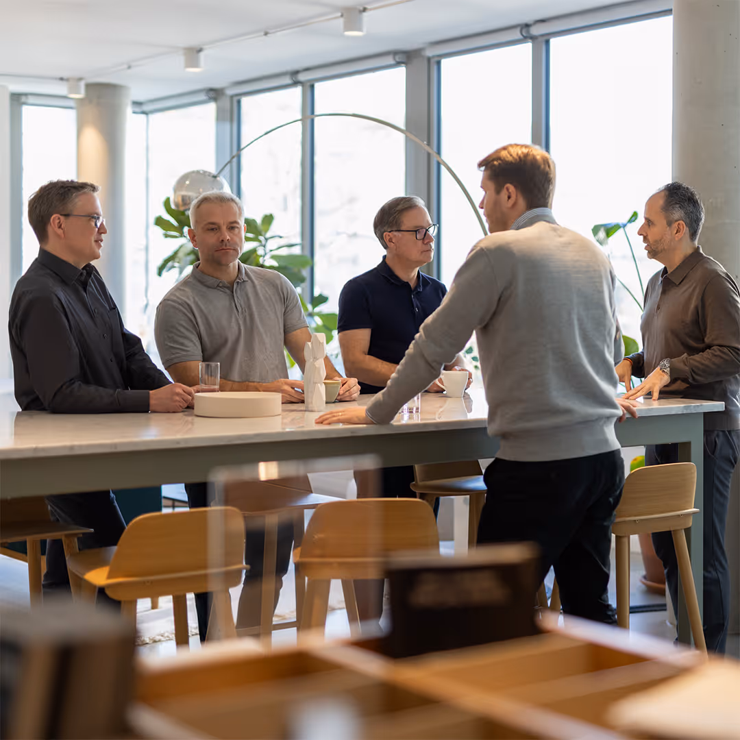 Five men having a discussion around a large table in a bright office with large windows and plants.