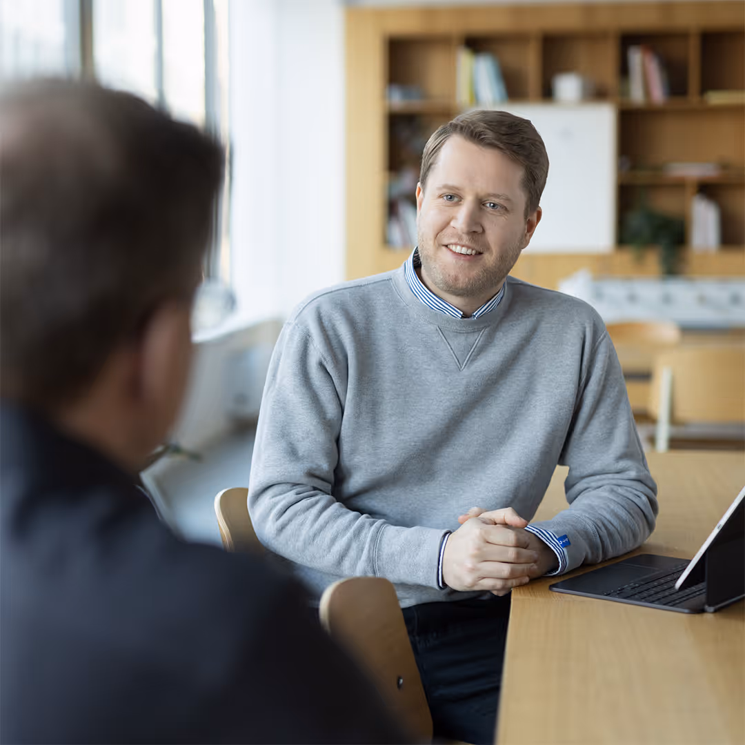 Man in a gray sweater smiling and talking to another person across a table with a tablet in a bright office space.