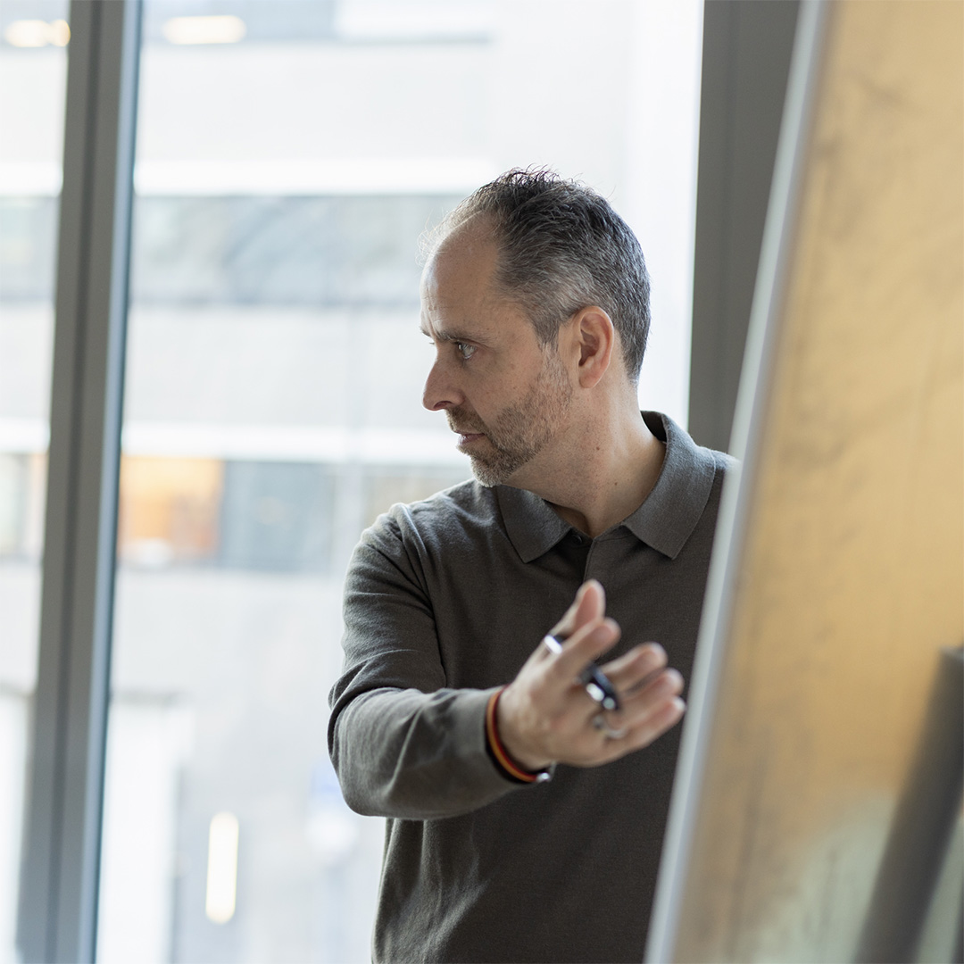Man with short hair and beard gesturing towards a board while looking to his left in a modern office room.