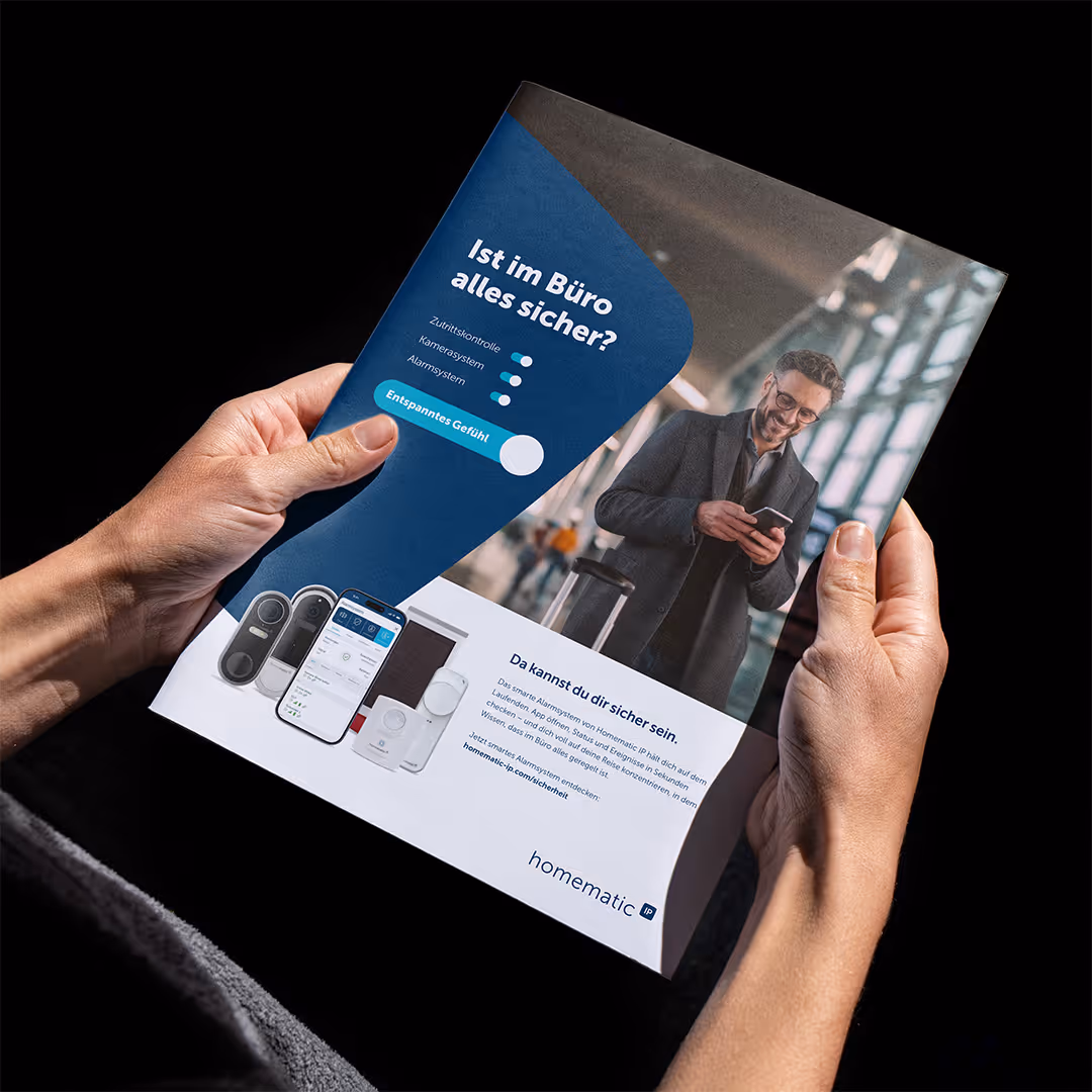 Hands holding a brochure with a man using a smartphone in an office setting, promoting Homematic IP office security system.