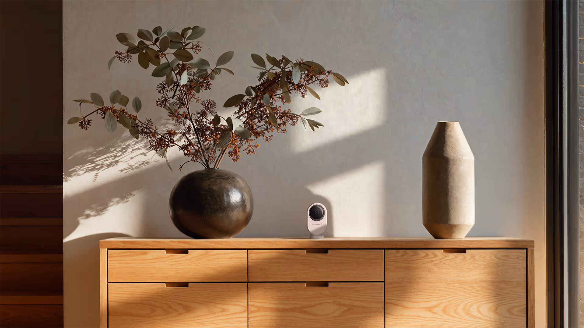 Modern wooden cabinet with a round dark vase holding dried branches on the left, a small security camera in the center, and a tall beige geometric vase on the right, under sunlight casting shadows on a plain wall.