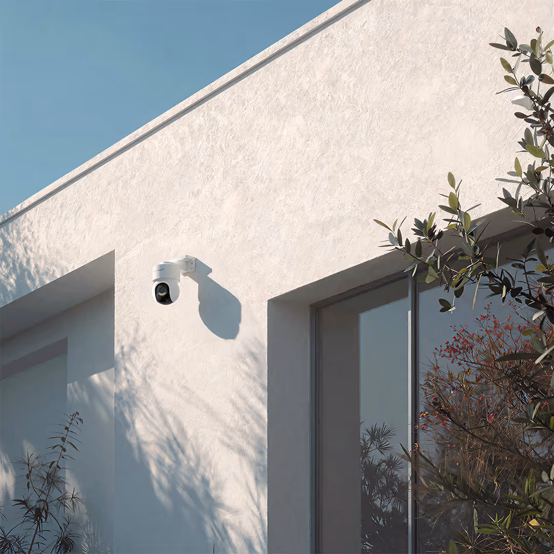 White outdoor security camera mounted on a white textured wall of a modern building beside a glass window and plants.