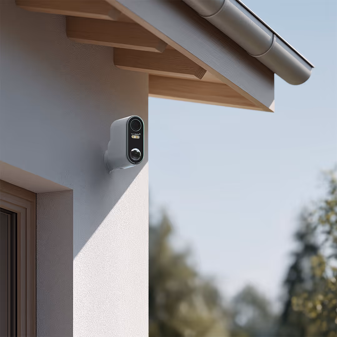 Outdoor security camera mounted on a gray wall under a wooden roof overhang with trees blurred in the background.